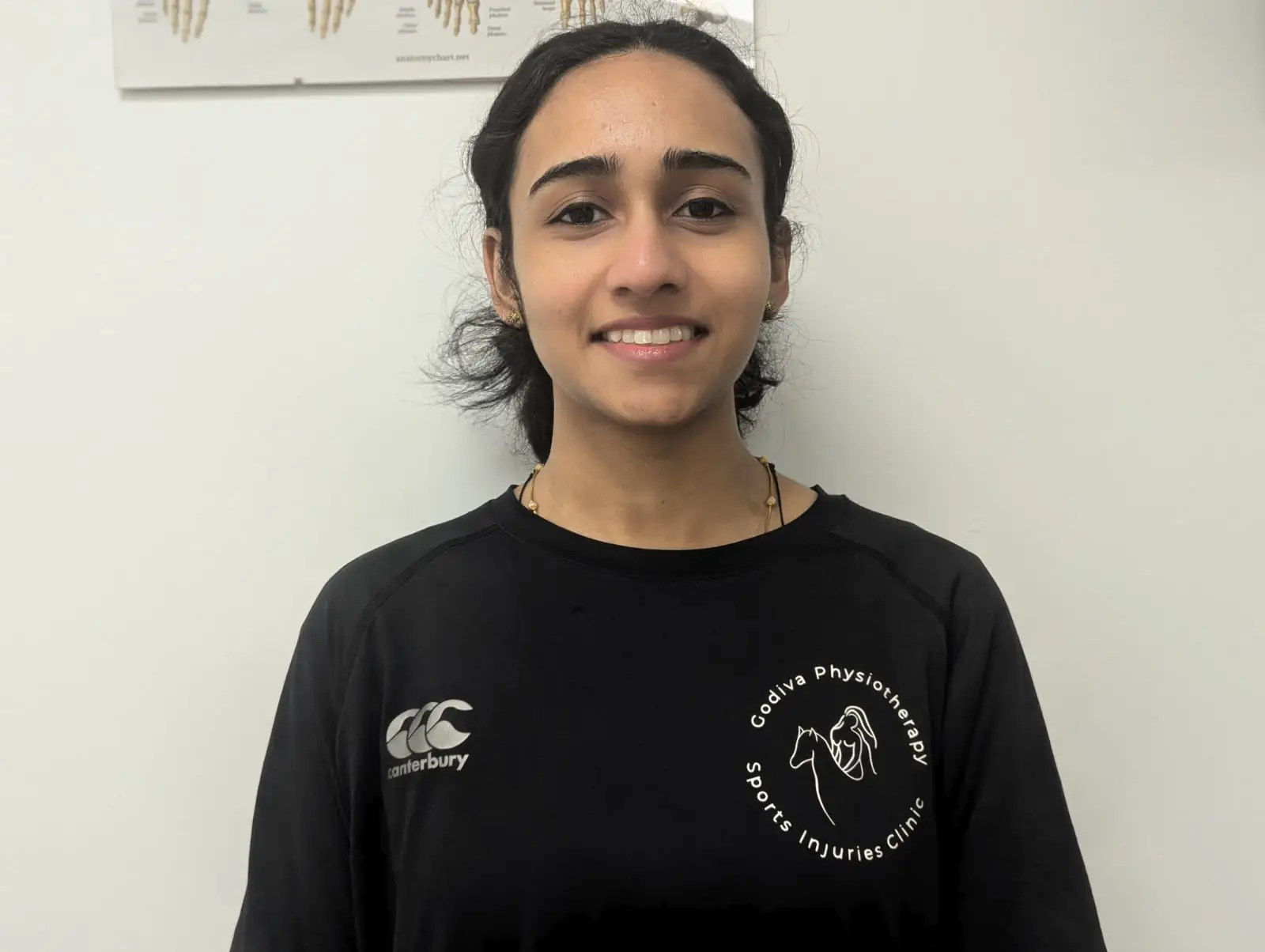 A person wearing a black sports shirt with a logo stands smiling in front of a white wall with a chart partially visible in the background.