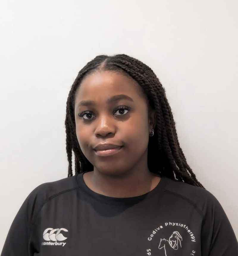 A person with long braids is wearing a black shirt with a logo. They are standing against a plain, light-colored background.