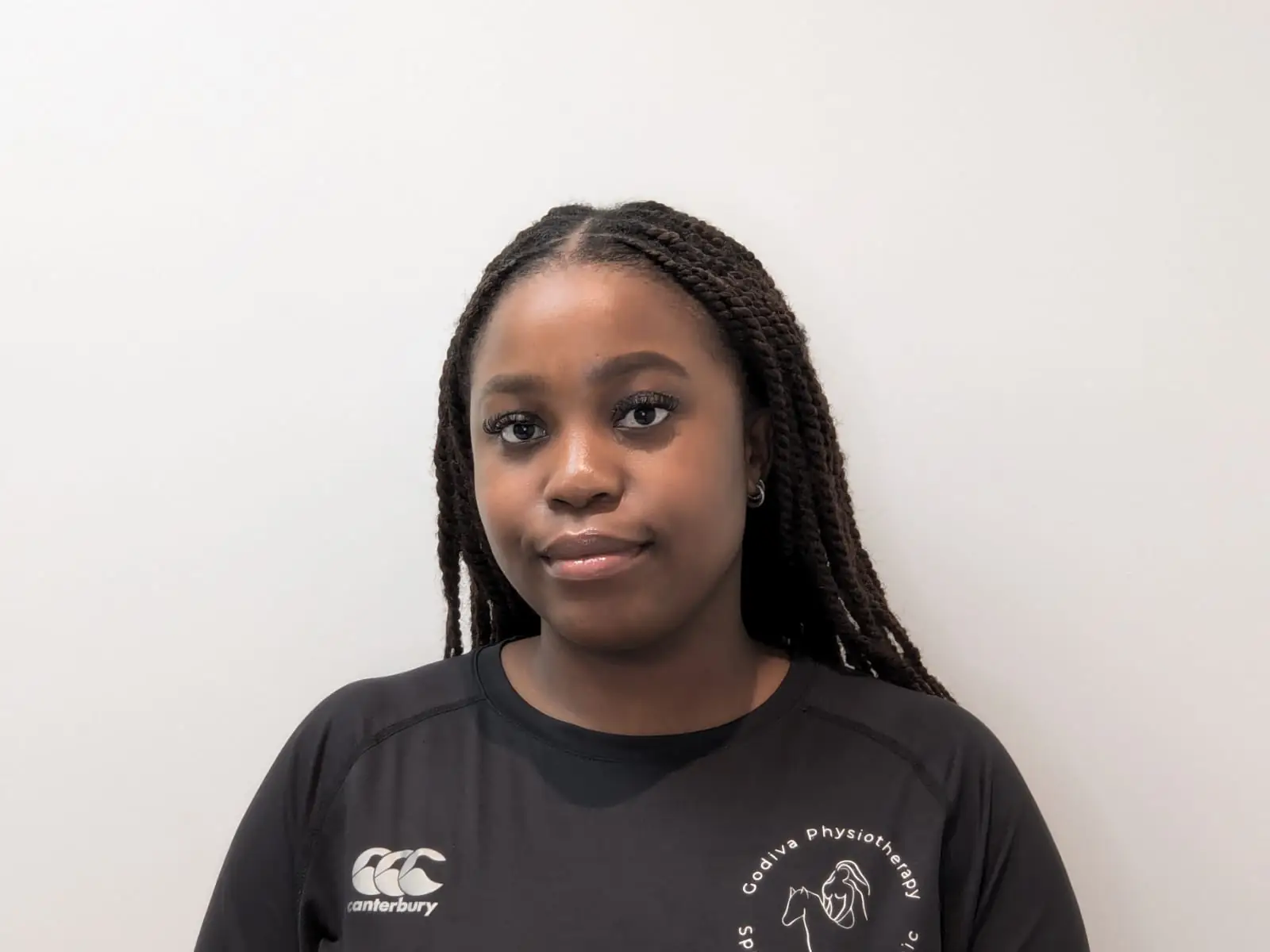 A person with long braids is wearing a black shirt with a logo. They are standing against a plain, light-colored background.