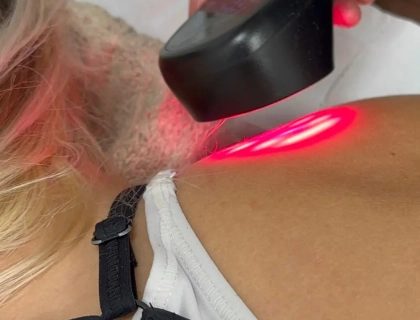 A person's shoulder is being treated with a handheld red light therapy device. The light is focused on the shoulder area, and the person is wearing a white top with black straps.