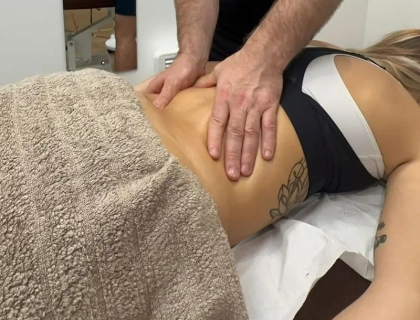 A person receiving a massage while lying on a table. The person's lower back and upper body are visible, with a towel covering the lower half. Hands of the masseuse are pressing on the back. The person has a tattoo on their side.