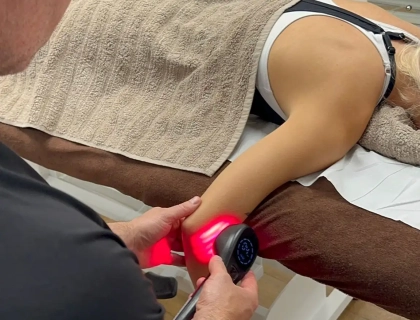 Person receiving a massage with a handheld massage gun on their arm in a therapy room. They are lying face down, covered with a beige towel. The therapist is focusing on the upper arm area, and the device emits a red light.