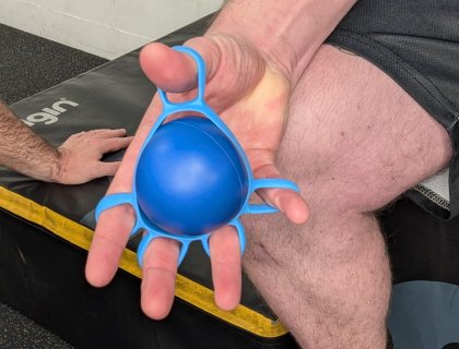 A person holding a blue hand exercise ball with a resistance band wrapped around their fingers. The person is seated on a padded surface in a gym setting, and another person is visible in the background.