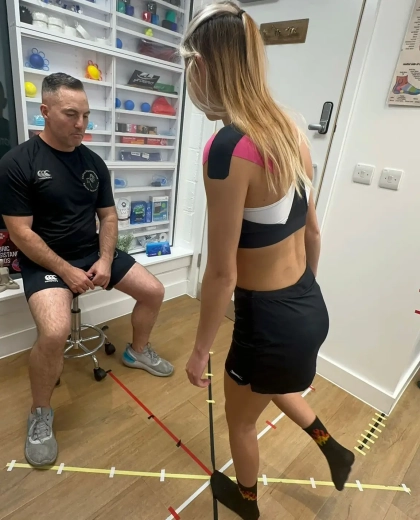 A woman performs a balance test on a taped pattern on the floor, watched by a man sitting on a stool. Shelves with various small items are visible in the background.