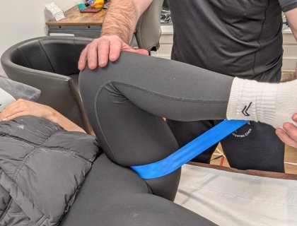 A person lies on a therapy table undergoing a physiotherapy session. One leg is bent and held by a therapist using a blue resistance band. The therapist supports the leg using hands, while the person wears black leggings and white socks.