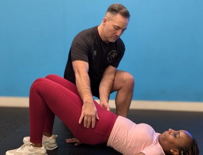 A fitness trainer guides a woman in performing a bridge exercise. She is lying on her back with knees bent, wearing pink leggings and a pastel top, while the trainer kneels beside her, offering support. The background is a solid blue wall.