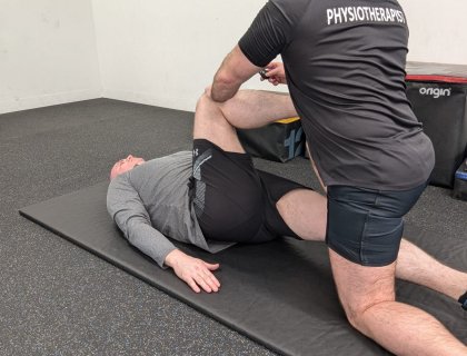 A physical therapist assists an older man lying on a mat, holding his bent leg during a stretching exercise. The therapist is wearing a shirt labeled "Physiotherapy.