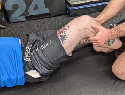 A person wearing a blue shirt and black shorts with tattoos is lying on the floor while another person, also with visible tattoos, supports their bent knee. The scene appears to be in a gym or physical therapy setting.