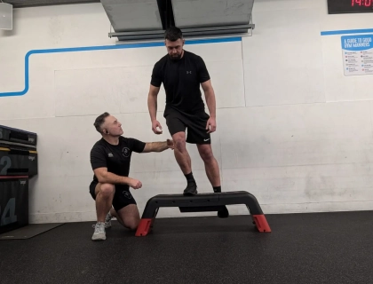 A man in a black shirt and shorts steps onto a platform as another man kneels beside him for support. Both are in a gym setting with storage boxes and a digital clock visible in the background.