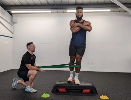 A person stands on a step platform with arms crossed and a resistance band around their knees, held by a trainer kneeling beside them. The setting is a gym with various colored cones on the floor.