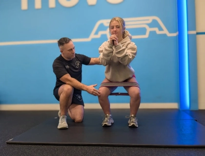 A coach guides a woman in performing a squat exercise, aiding her hamstring rehabilitation. She has a resistance band around her thighs, wearing a beige hoodie and gray sneakers. The setting is a gym with a blue background and the word "MOVE" partially visible.