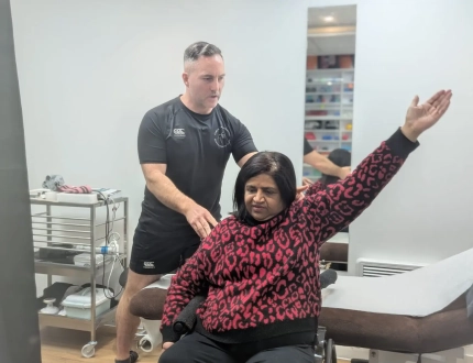 A physical therapist assists a woman in a wheelchair, focusing on shoulder treatment as she raises her left arm. The room, equipped with a massage table and various supplies, provides an ideal setting for alleviating shoulder pain.