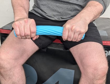 A person sitting on a workout bench, holding a blue resistance bar with both hands. They are wearing a gray shirt and black shorts. The background features a padded exercise surface.